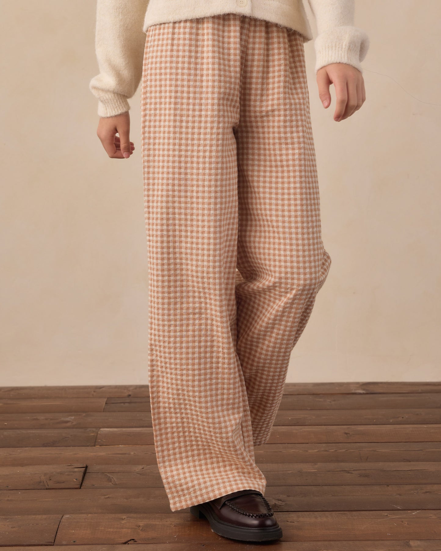 A person wearing loose, wide-leg trousers in a beige and brown gingham check pattern, styled with a cream knit sweater and dark brown leather loafers, standing on a wooden floor against a neutral beige wall.