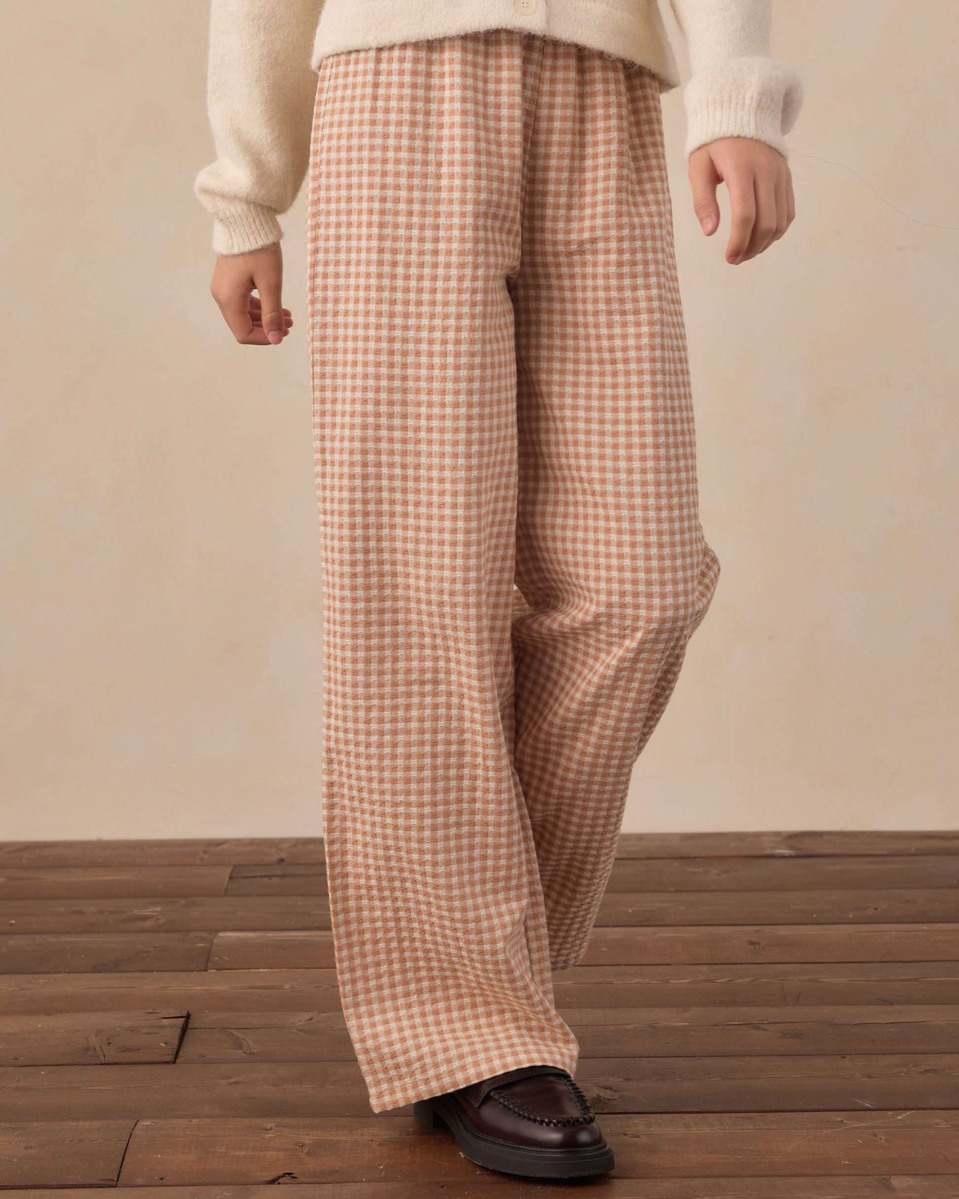 A person wearing loose, wide-leg trousers in a beige and brown gingham check pattern, styled with a cream knit sweater and dark brown leather loafers, standing on a wooden floor against a neutral beige wall.