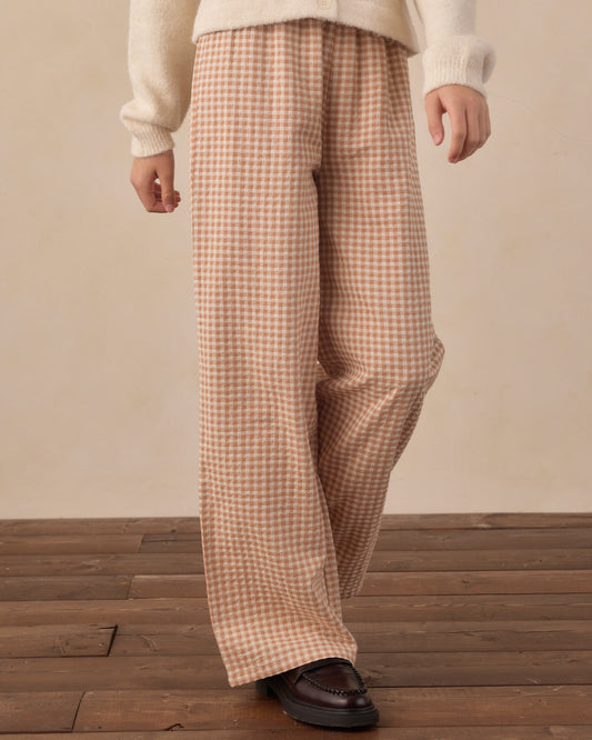 A person wearing loose, wide-leg trousers in a beige and brown gingham check pattern, styled with a cream knit sweater and dark brown leather loafers, standing on a wooden floor against a neutral beige wall.