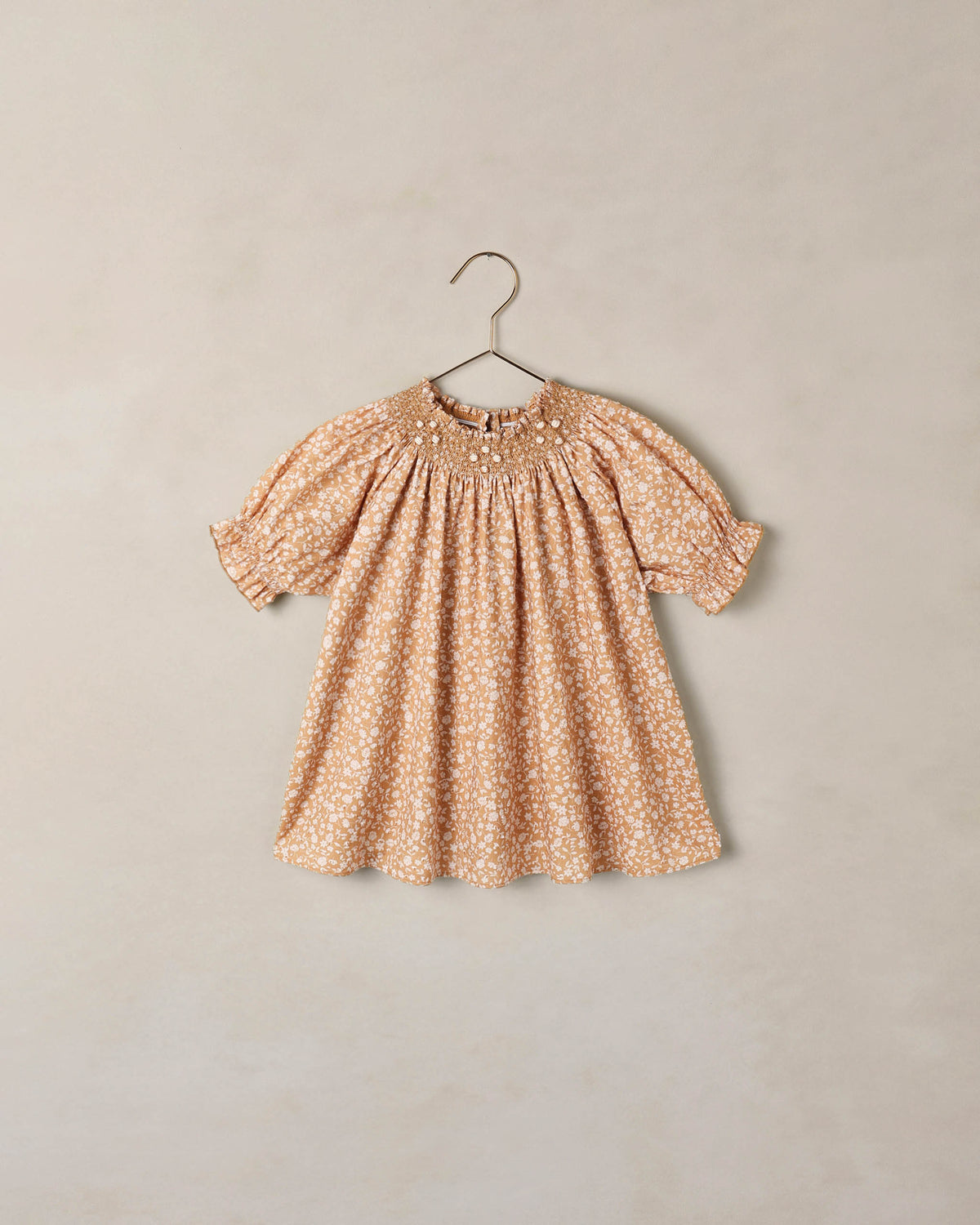 A short-sleeve baby dress with a delicate white floral pattern on a warm peach-beige background, hanging on a metal hanger against a textured beige wall.