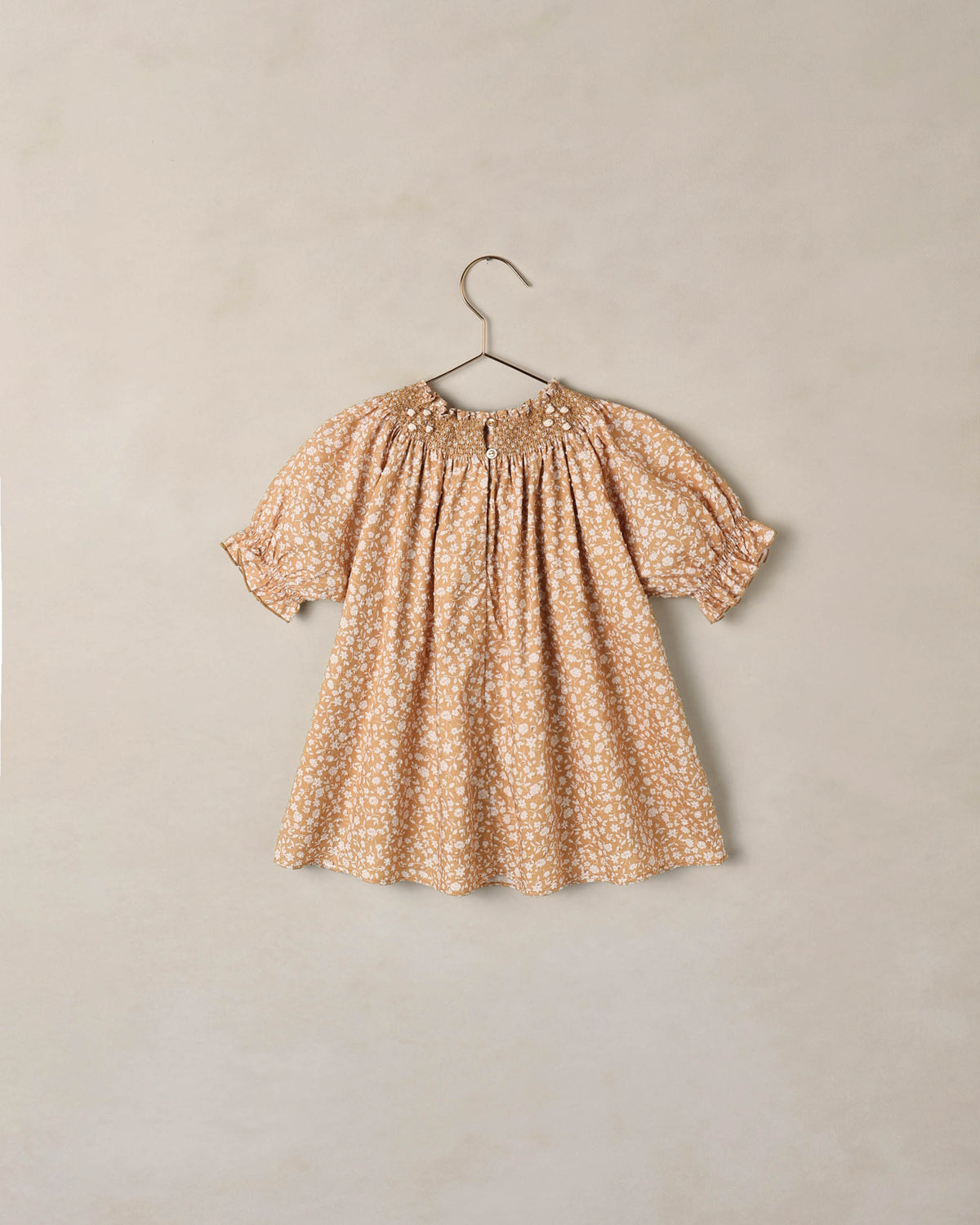 A short-sleeve baby dress with a beige and white floral pattern, featuring a smocked bodice and puff sleeves, hanging on a metal hanger against a textured beige wall.