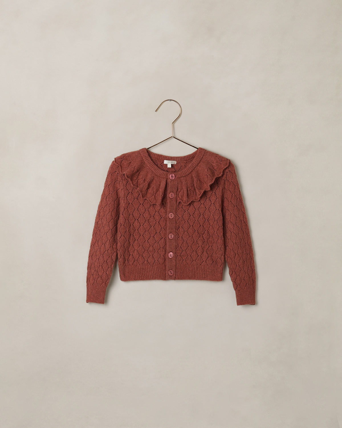 A terracotta-colored cable-knit cardigan sweater with a ruffled collar and button-front closure, hanging on a simple metal hanger against a textured beige wall.