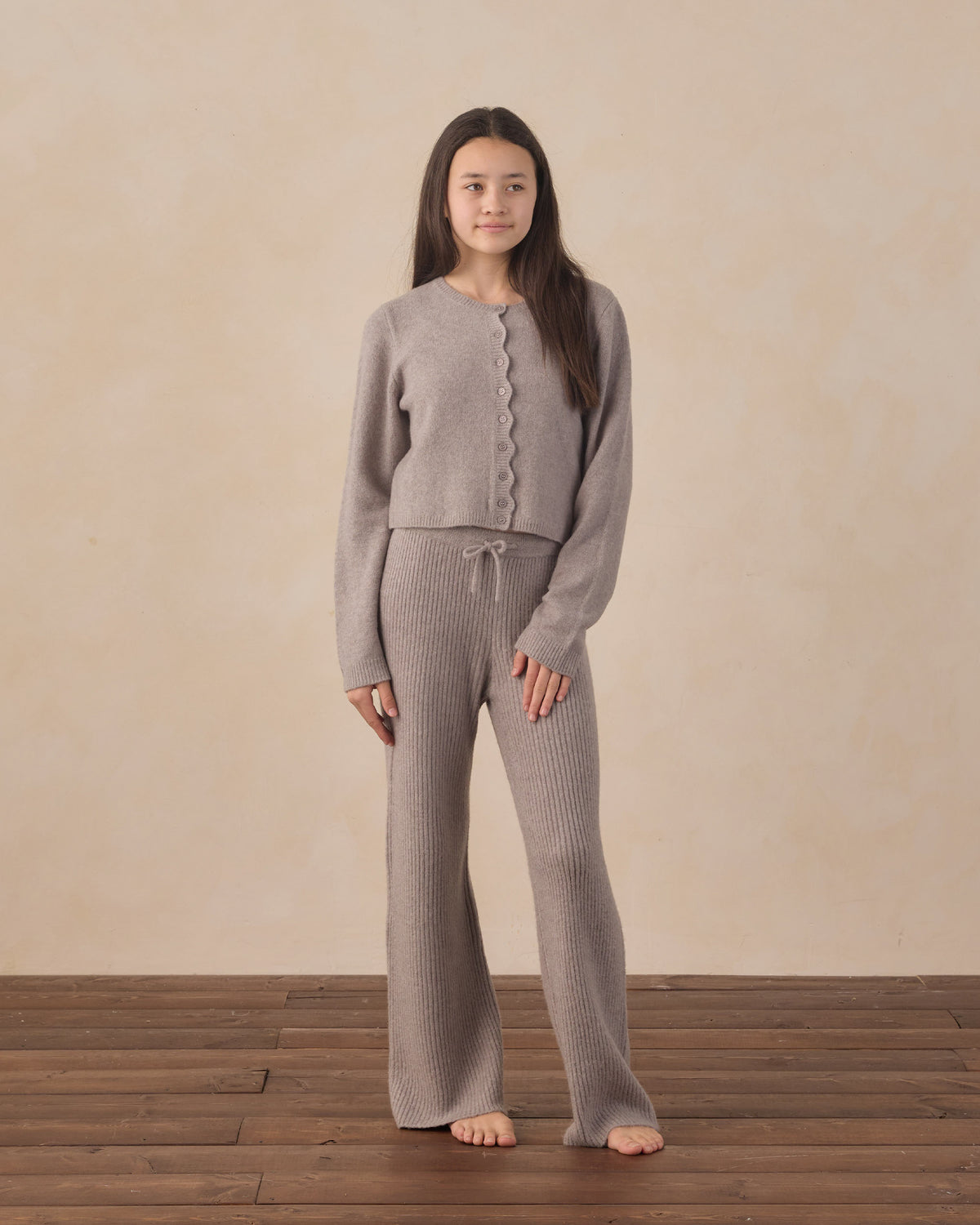 Woman wearing a gray knit outfit standing on a wooden floor with a beige wall background