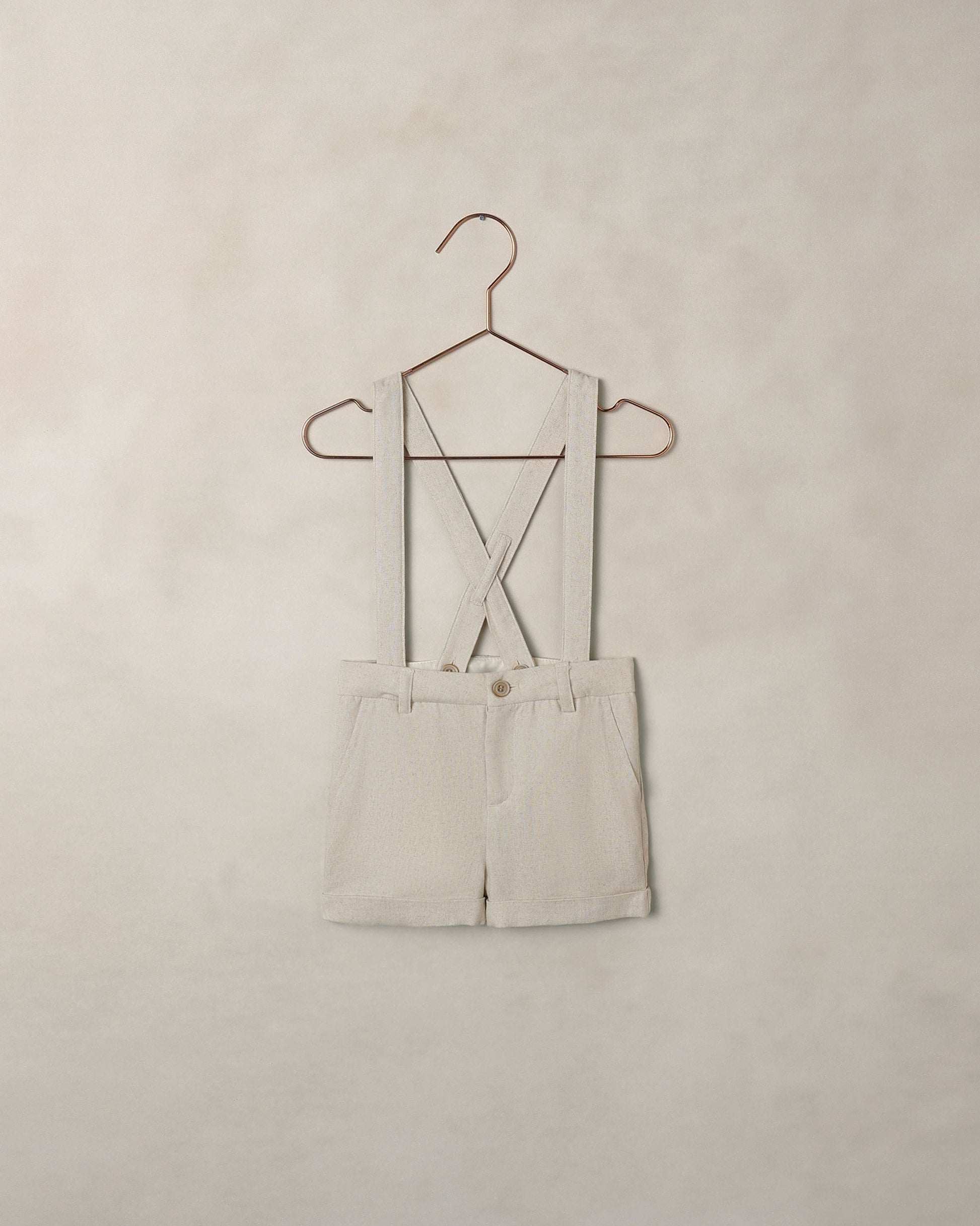 Beige shorts overalls on a hanger against a beige wall