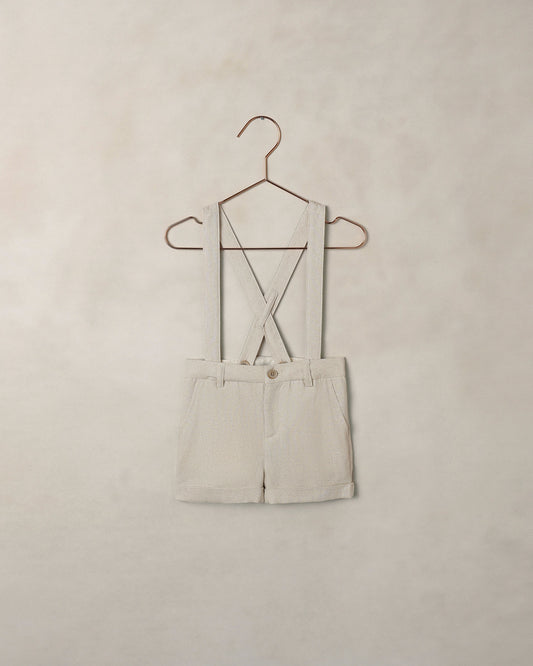 Beige shorts overalls on a hanger against a beige wall